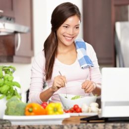 Cooking woman looking at computer while preparing food in kitchen. Beautiful young multiracial woman reading cooking recipe or watching show while making salad.